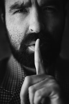 photo en noir et blanc du visage d'un homme barbu avec l'index à la verticale devant la bouche (demande de silence